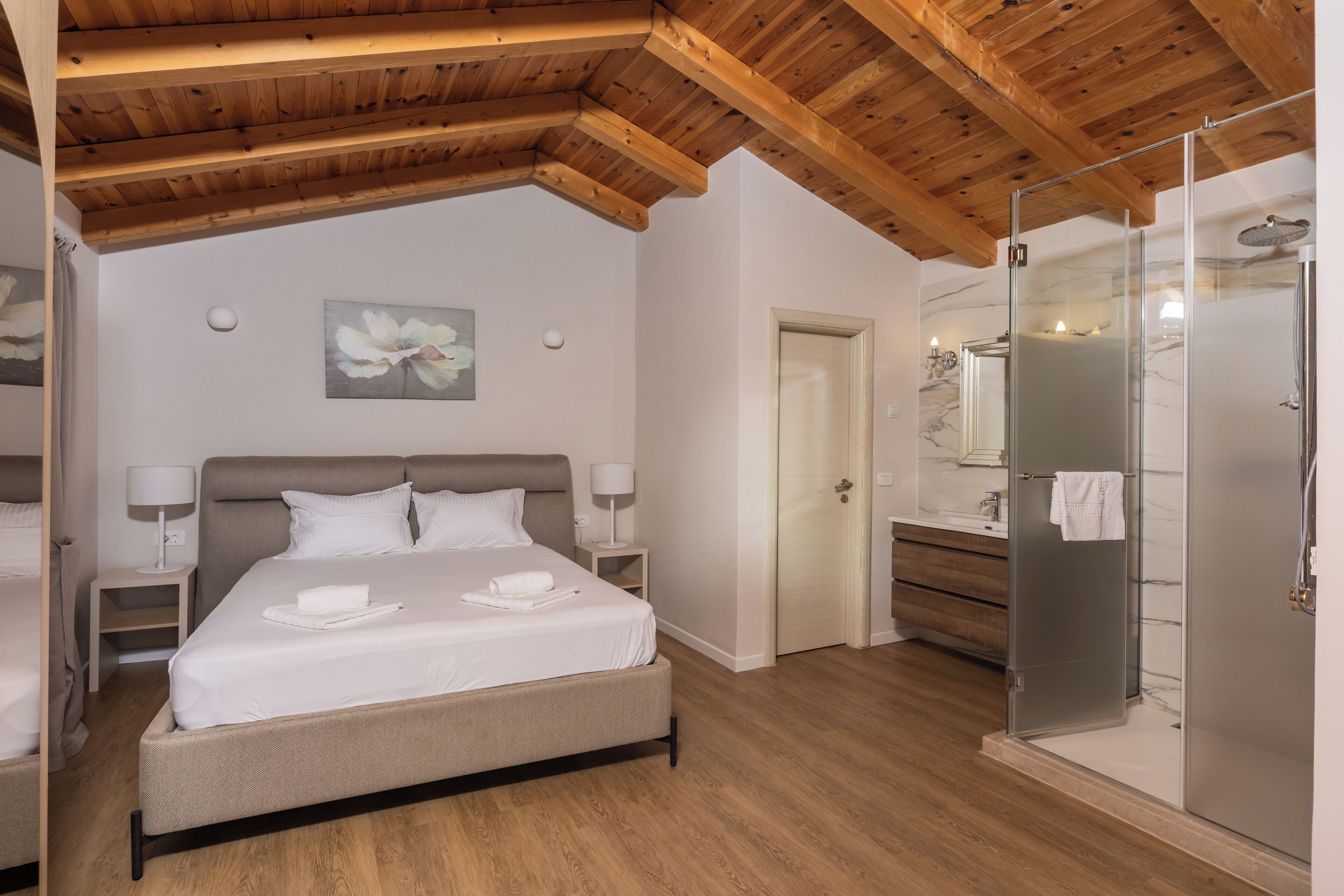 A bedroom with a bed, bedside tables, a nightstand, a wooden ceiling, a bathroom with a shower, a mirror, and a door.