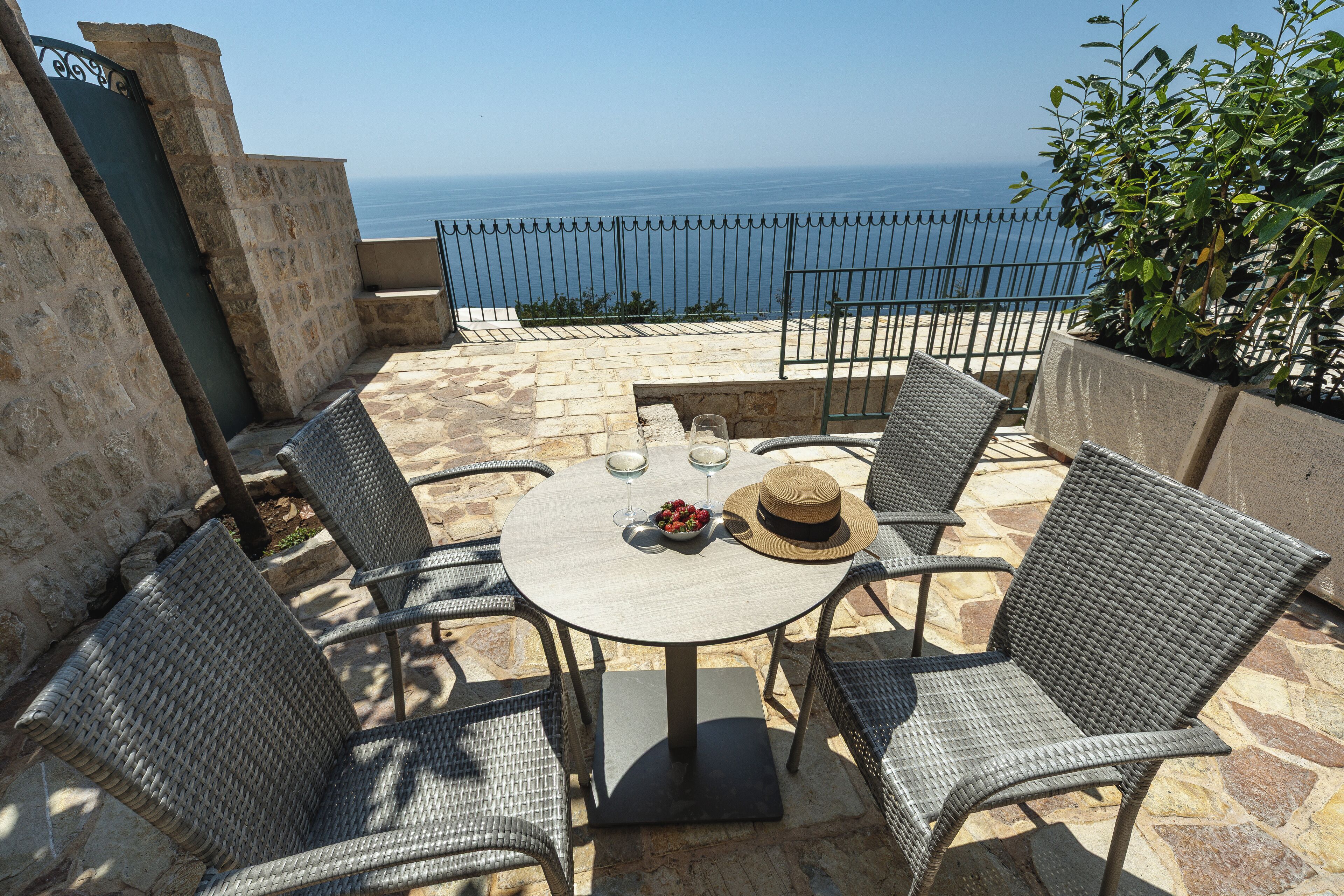 A round table with wicker chairs, a hat, and a bowl of fruit on a patio overlooking the sea.