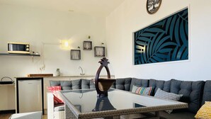 Un salon moderne avec un canapé gris, une table basse en verre et un fauteuil en osier. On y trouve également un petit coin cuisine équipé d’un réfrigérateur et d’un four à micro-ondes. Un élément décoratif mural et des tableaux encadrés sont visibles.