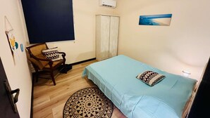 Une petite chambre à coucher accueillante, avec un lit simple, une chaise, un tapis rond à motifs, une fenêtre avec un store foncé, un luminaire au plafond et un climatiseur fixé au mur.