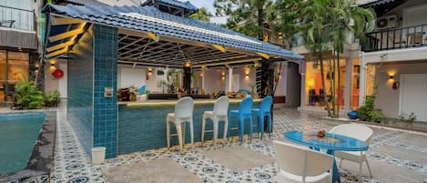 Et poolområde med flisegulv, et overdækket loungeområde med stole og en bar.