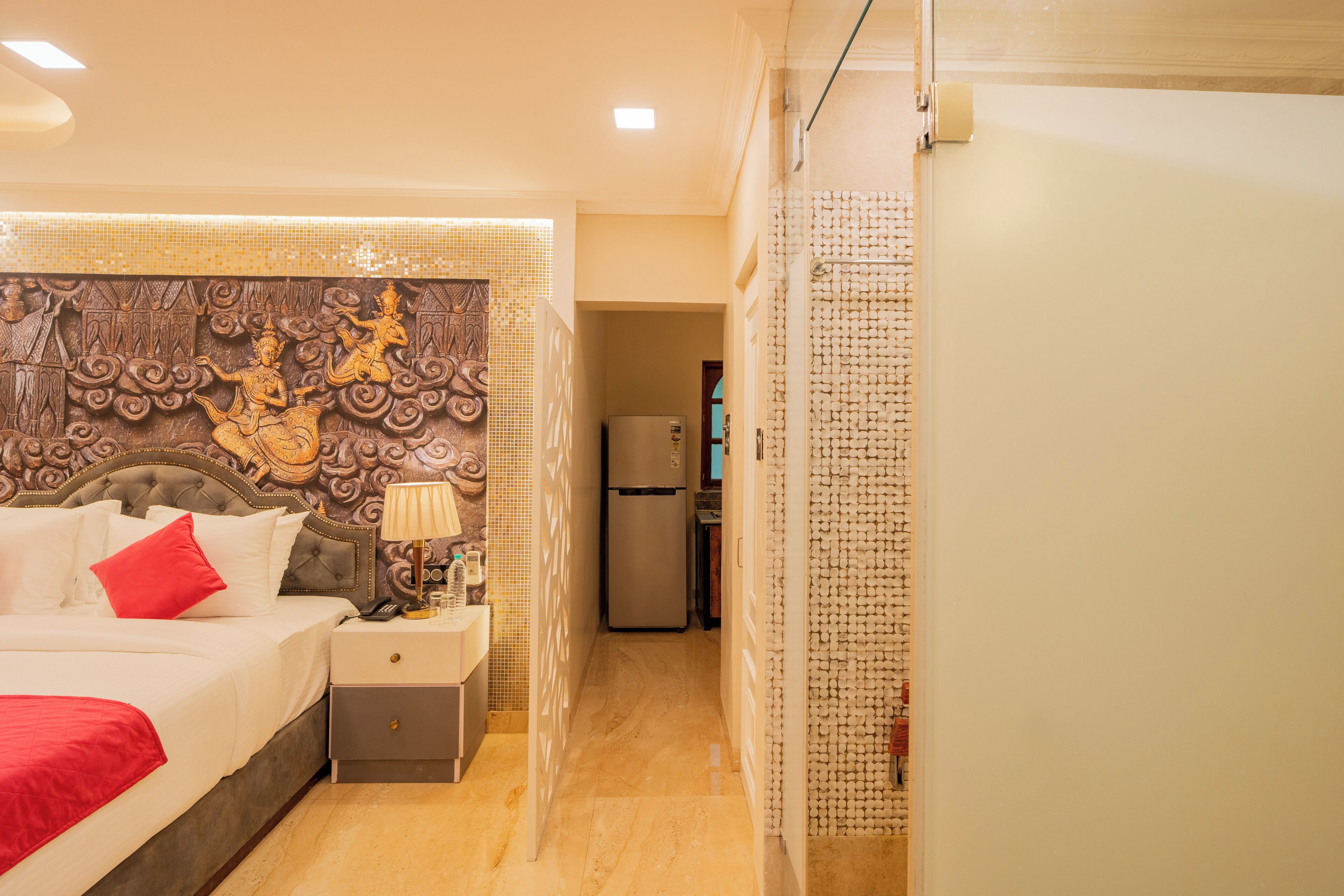A hotel room with a large bed, a nightstand, a wall with intricate carvings, a refrigerator, and a bathroom with a shower curtain.