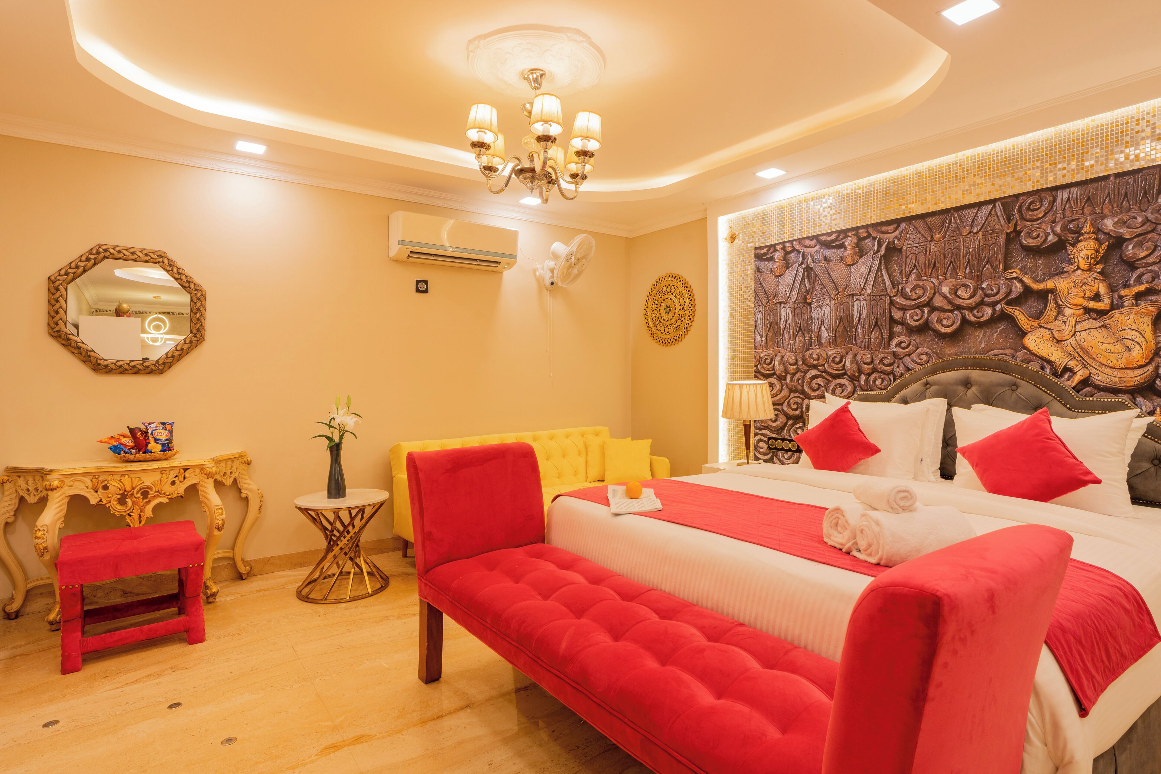 A hotel room with a large bed, a red bench, a small table, and a decorative headboard.