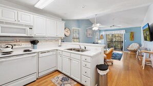 Una cocina moderna con muebles blancos, microondas, estufa y lavabo. Hay un comedor con una silla y una mesa, y un balcón con vistas.