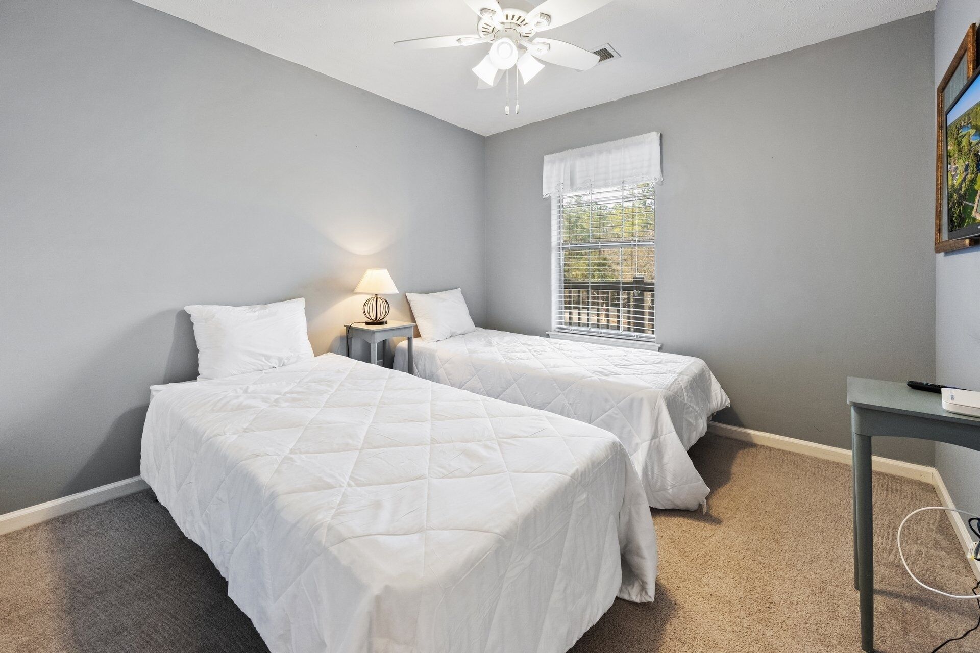 A bedroom with two beds, a ceiling fan, a window with blinds, and a desk with a lamp.
