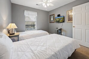 A bedroom with two beds, a ceiling fan, a window with blinds, a TV, and framed pictures on the wall.