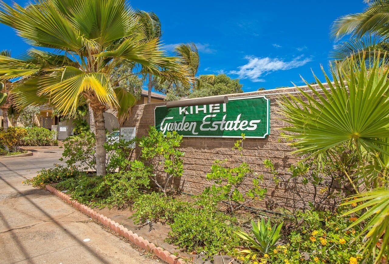 A sign for KIHEI Garden Estates with a brick wall and tropical plants.