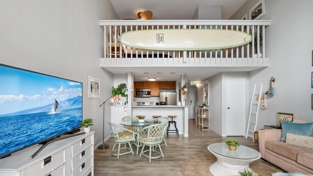 Amplia sala de estar con un televisor de pantalla plana grande, una mesa de comedor con sillas, una mesa redonda de centro y un sofá. Se ve una cocina con electrodomésticos modernos al fondo.