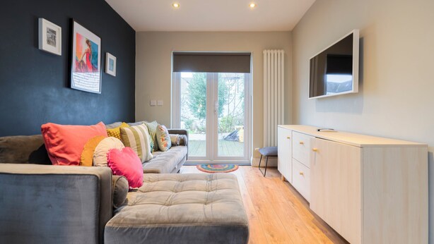 Un salon moderne avec un canapé gris, des coussins colorés, un téléviseur à écran plat fixé au mur et une porte-fenêtre coulissante donnant sur un espace extérieur.