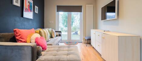 Un salon moderne avec un canapé gris, des coussins colorés, un téléviseur à écran plat fixé au mur et une porte-fenêtre coulissante donnant sur un espace extérieur.