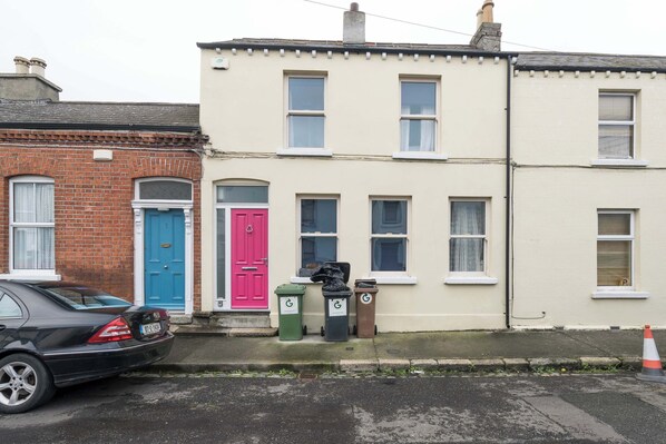A row of houses with distinct colored doors, a parked car, and three trash bins.