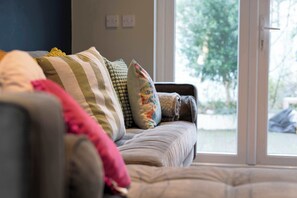 A sofa with multiple cushions, a window with a view of greenery, and a wall switch.