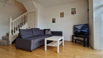 Una sala de estar con un sofá gris, una mesa de centro blanca, un televisor sobre un mueble bajo, una escalera y un ventanal con cortinas.