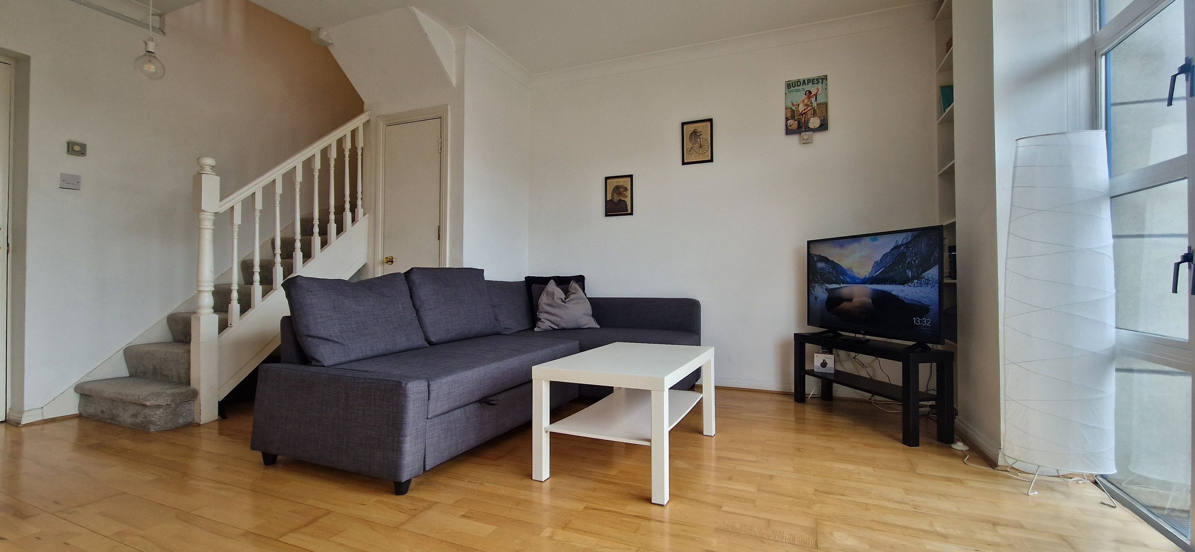 Una sala de estar con un sofá gris, una mesa de centro blanca, un televisor sobre un mueble bajo, una escalera y un ventanal con cortinas.