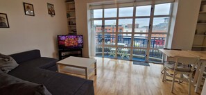 A living room with a sofa, a coffee table, a TV, and a balcony with a view of buildings.