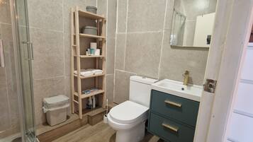 Un baño con inodoro, lavamanos, espejo, repisa con artículos de aseo y cubo de basura.