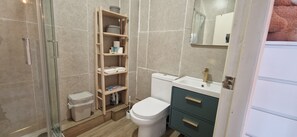 Un baño con inodoro, lavamanos, espejo, repisa con artículos de aseo y cubo de basura.