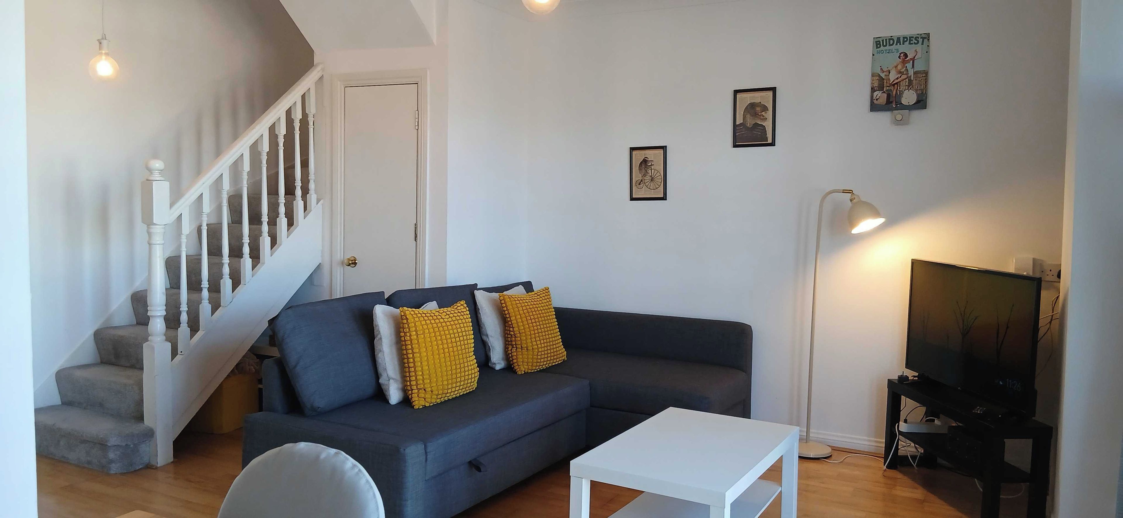 Una sala de estar con un sofá gris oscuro, dos almohadas amarillas, una mesa de centro blanca, un mueble de televisión con televisor, y una escalera con barandillas blancas.