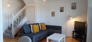 A living room with a dark grey sofa, two yellow pillows, a white coffee table, a TV stand with a TV, and a staircase with white railings. - Waterfront 2BD Flat with Balcony, Central Dublin (Dublin)