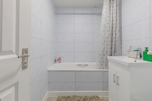 A bathroom with a white bathtub, a shower curtain, and a vanity with toiletries. - Riverside 2BD Flat, Short Walk to the ExCel Centre (Greater London)