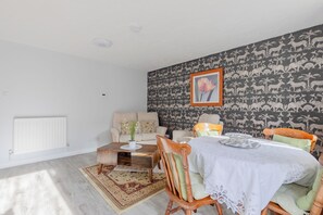 A living room with a dining area, white walls, a patterned wallpaper, a radiator, a wooden coffee table, a sofa, and a dining table with chairs. - Riverside 2BD Flat, Short Walk to the ExCel Centre (Greater London)