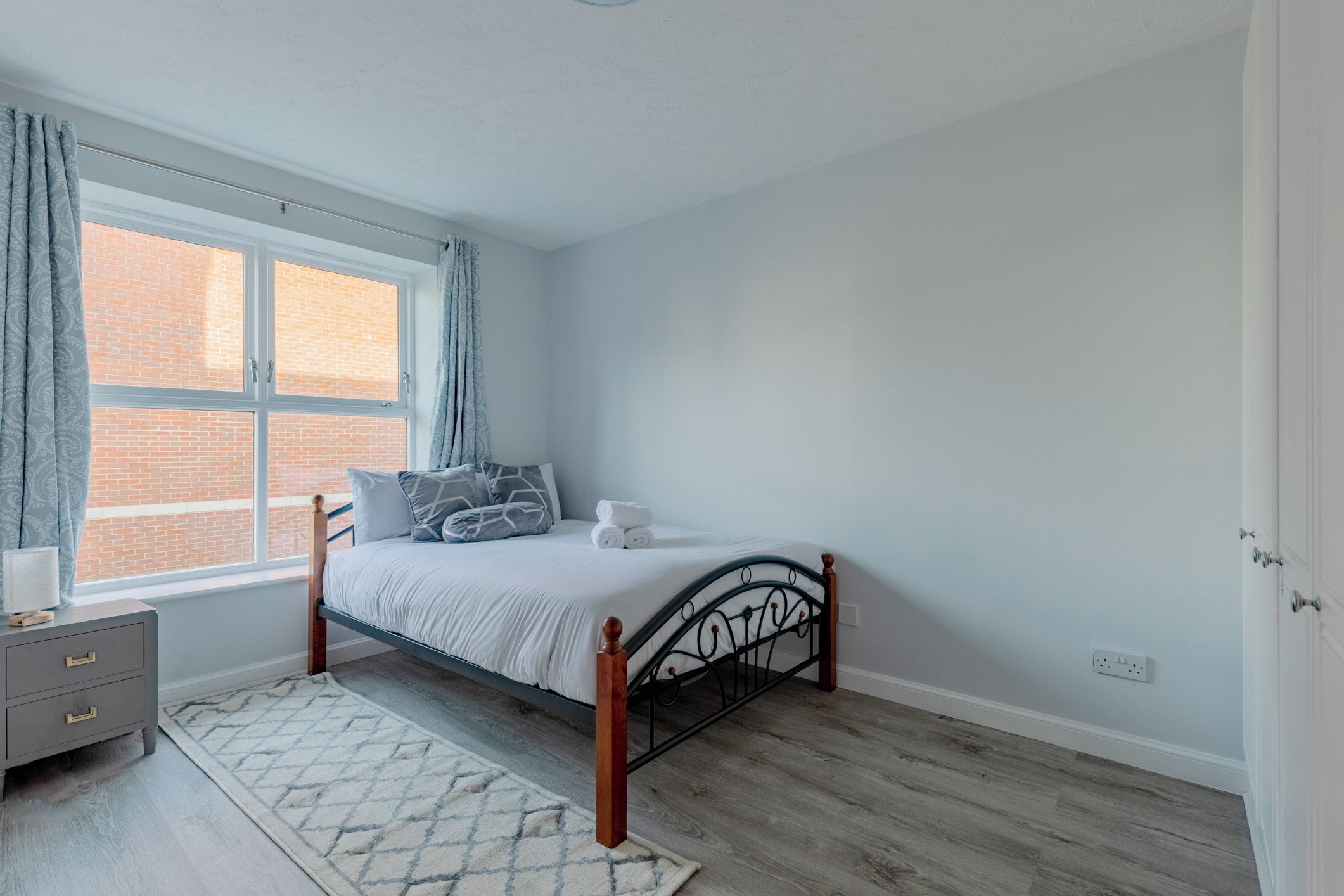 A bedroom with a bed, a nightstand, a window with curtains, and a view of a brick building.