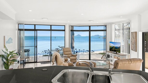 A modern kitchen with a large island, a dining area with a view of the sea, and a living area with a television and artwork.