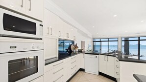 A modern kitchen with white cabinetry, a built-in oven, and a dishwasher. It features a large window with a view of the sea.