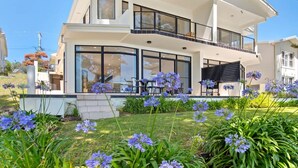 A modern two-story house with large windows, a balcony, and a well-maintained garden with purple flowers.