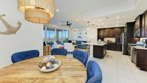 A modern kitchen with dark wood cabinets, a central island, and a dining area with a wooden table and blue chairs. - Anglers Bay at One Particular Harbour 392-103 · Lagoon Views · 5-Night Min (Bradenton)