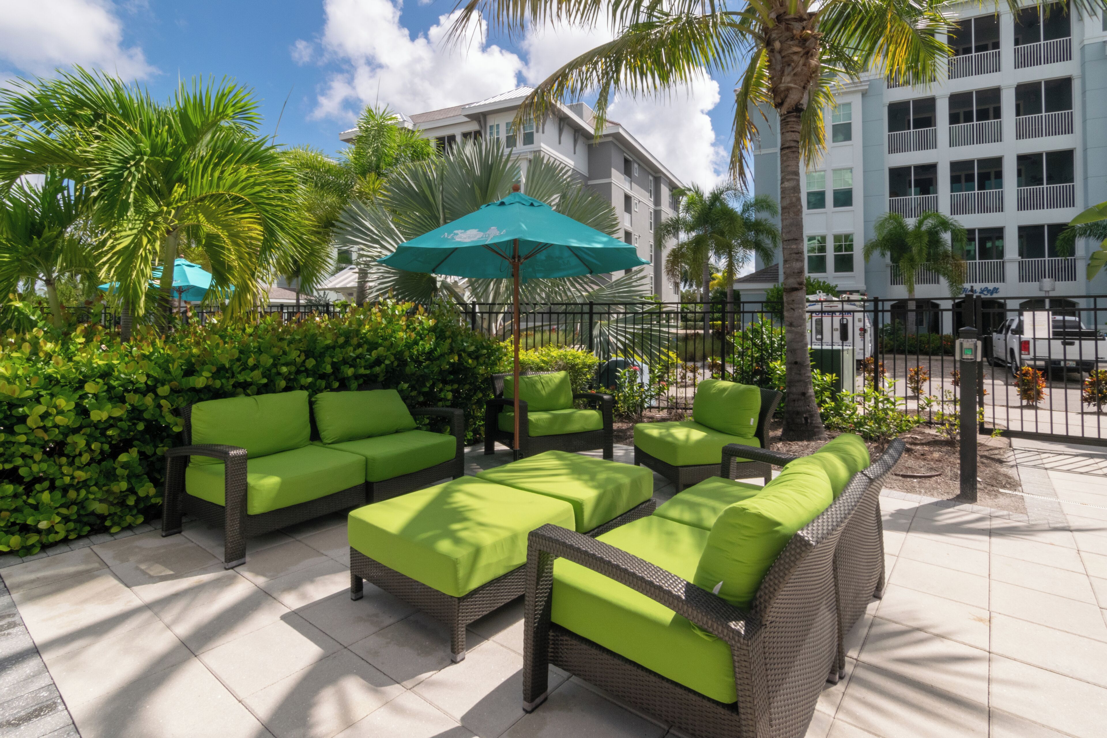 An outdoor seating area with wicker furniture and green cushions, surrounded by palm trees and a building in the background.