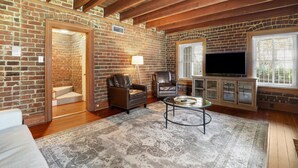 A cozy living room with exposed brick walls, wooden beams, a fireplace, leather chairs, a TV, and a glass coffee table.