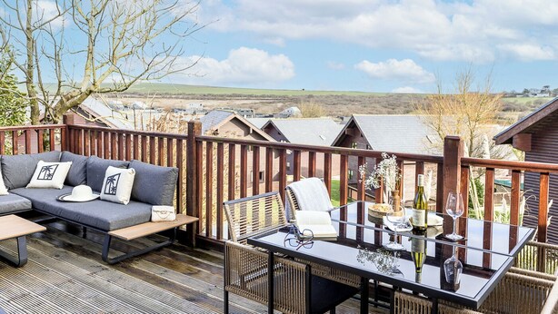 A deck with a sofa, chairs, and a table set for dining, overlooking a village with houses and hills. - The Palm Tree Lodge | Spacious contemporary 2 bed (Saint Columb Major)