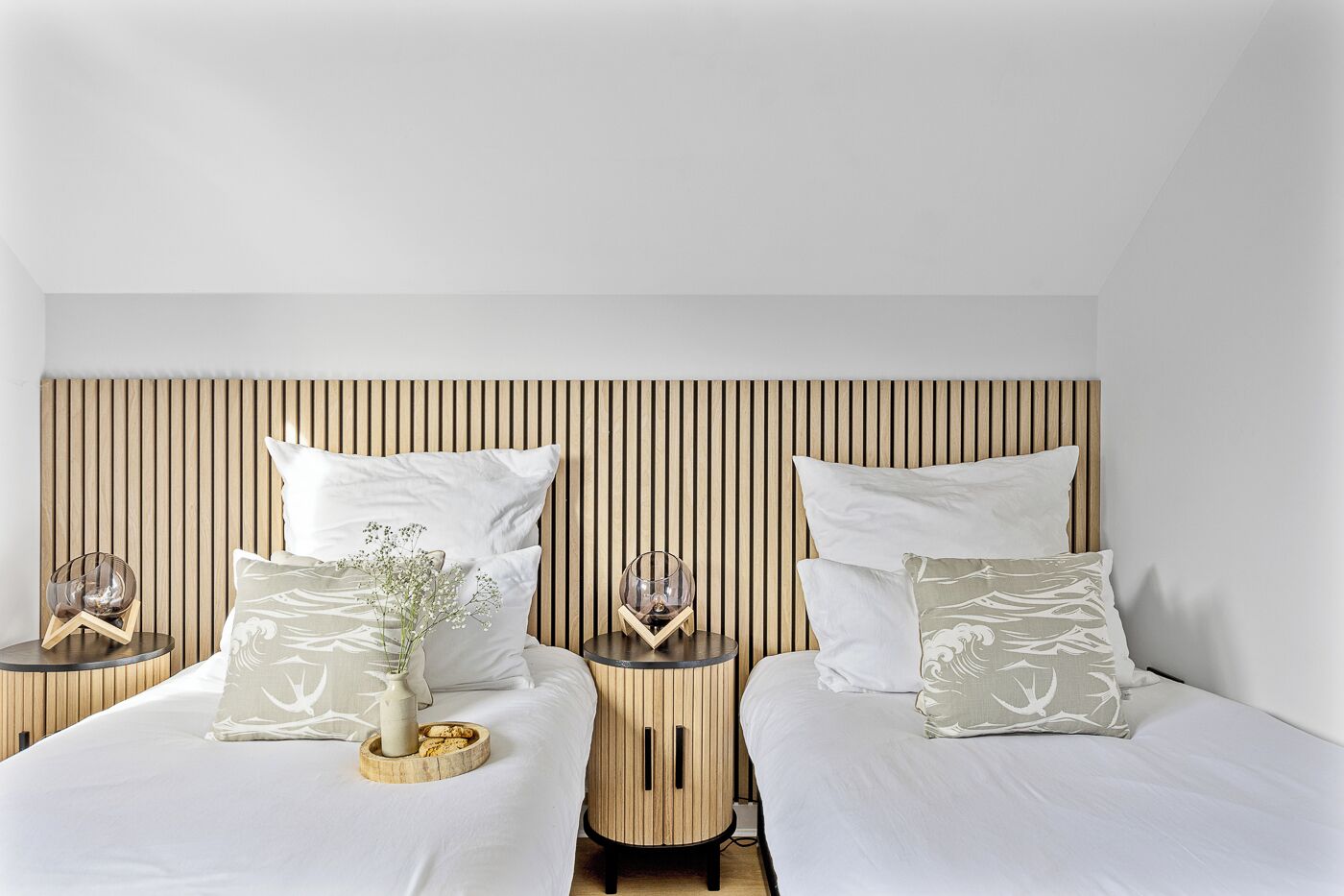 A modern bedroom with two beds, each with a wooden headboard, white bedding, and decorative pillows. There are bedside tables with glass lamps and a vase with flowers.