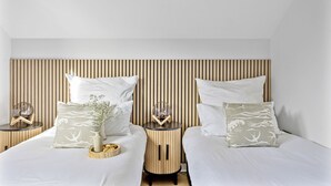 A modern bedroom with two beds, each with a wooden headboard, white bedding, and decorative pillows. There are bedside tables with glass lamps and a vase with flowers. - The Palm Tree Lodge | Spacious contemporary 2 bed (Saint Columb Major)