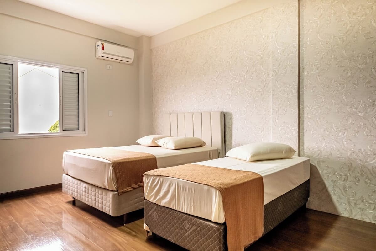 A room with two beds, a window with blinds, an air conditioning unit, and patterned wallpaper.