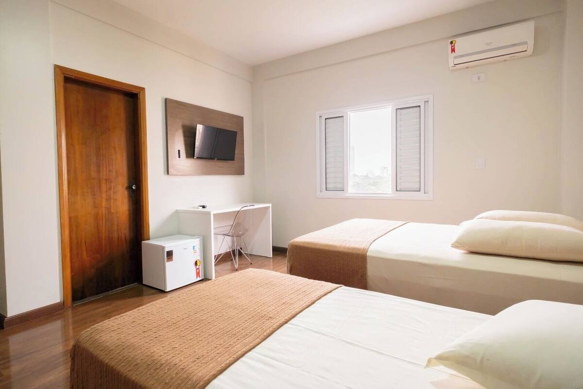 A hotel room with two beds, a desk, a chair, a TV, and a window with shutters.