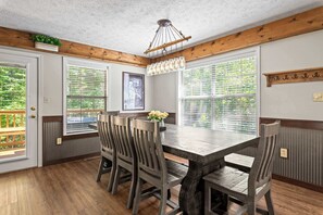 A dining area with a wooden table and chairs, large windows with blinds, a hanging light fixture, and a wooden ceiling.