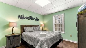 Een slaapkamer met een bed, twee bedlampen, een nachtkastje, een raam met jaloezieën en een decoratie aan de muur.