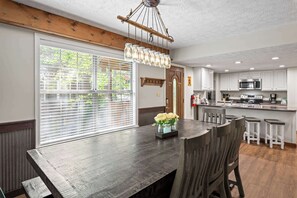 A dining area with a dark wooden table, white chairs, a hanging light fixture, and a window with blinds.
