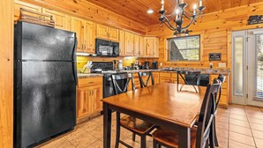 Interior de una cabaña de madera con cocina, una mesa de comedor, sillas, refrigerador, microondas y una lámpara de araña.