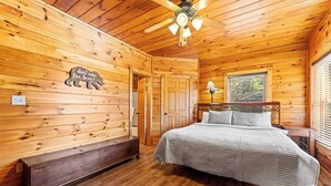 Dormitorio de cabaña de madera con cama, banco, ventilador de techo y aplique de pared.