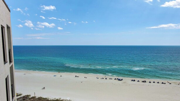 A beach with clear blue water and a white sandy shore. There are people and umbrellas on the beach. - 🌴🌞🌴Beachfront Condo | Seasonally Heated Pool | Navarre Towers 1205🌴🌞🌴 (Navarre Beach)