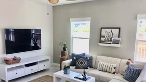 A modern living room with a flat-screen TV, a white sofa, a coffee table, and a large window providing natural light.