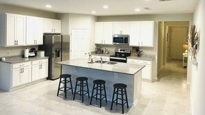 A modern kitchen with white cabinets, a large island with bar stools, stainless steel appliances, and a tiled floor.