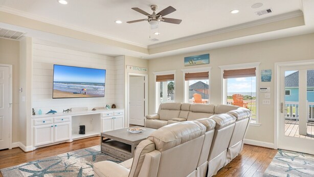 A spacious living room with a large beige sectional sofa, a flat-screen TV mounted on the wall, and a ceiling fan.