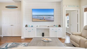 A living room with a large TV displaying a beach scene, a white cabinet with decorative items, a beige sofa, and a sign indicating a beach direction.
