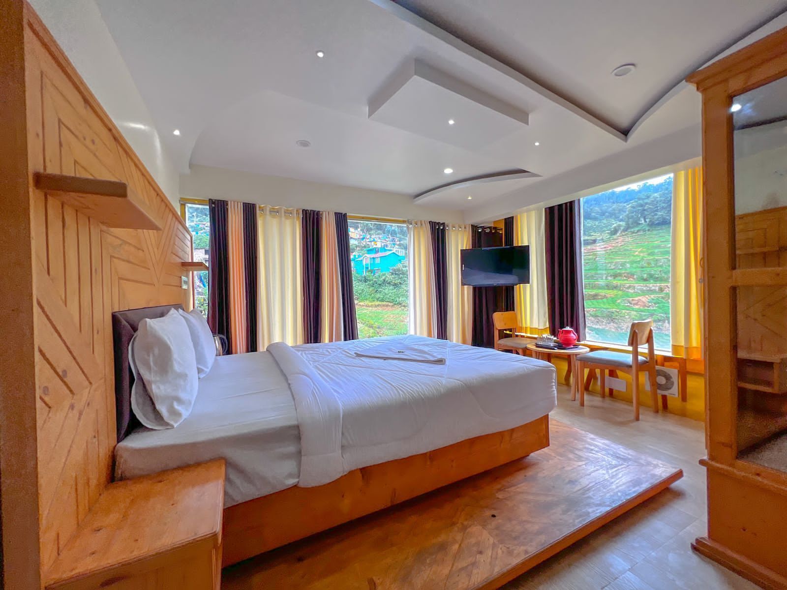 Une chambre avec un grand lit, une télévision, une petite table sur laquelle se trouve un pot rouge, et une vue sur l’extérieur.