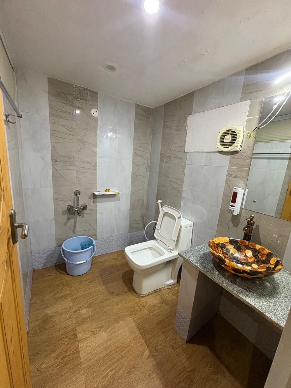 Un bagno con il water, un secchio blu, una parete doccia in marmo, un lavabo decorativo e una ventola a soffitto.