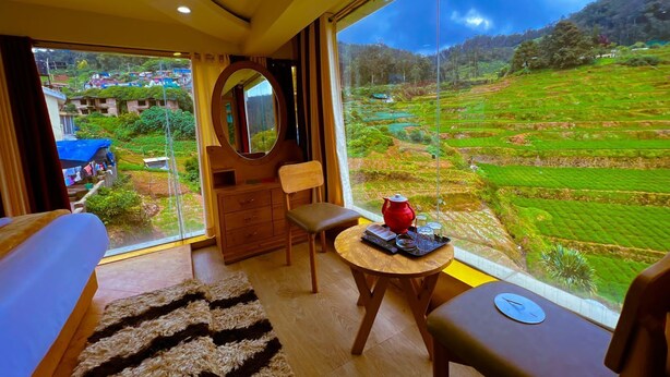 A room with a large window offering a view of terraced fields. There is a bed, a chair, a small table with a teapot, and a mirror. - Rozana Retreat (Ootacamund)
